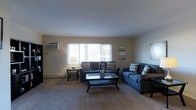 This single-floor space offers a functional and well-appointed layout designed for comfortable everyday living. The living area serves as the centerpiece of the home, featuring plush wall-to-wall carpeting that adds warmth underfoot and contributes to the overall cozy ambiance. A large window spans the exterior wall, allowing an abundance of natural light to fill the room while offering views of the surrounding greenery outdoors. The neutral-toned walls provide a versatile backdrop, complementing a range of furnishing styles. The living room is generously sized, comfortably accommodating two full sofas in a coordinated blue upholstery paired with chevron-patterned accent pillows, centered around a sleek glass-top coffee table. A wall-mounted HVAC unit and a baseboard heating system ensure year-round climate comfort. Subtle decorative touches, including wall art and a classic table lamp, contribute to the space's inviting character. The smooth ceiling and clean lines throughout the room reflect a straightforward, practical design sensibility. Overall, this space presents a livable and approachable environment with ample room for relaxation and everyday use.
