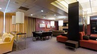 This single-floor commercial lounge space is thoughtfully designed to accommodate both casual gatherings and functional meetings. The interior is divided into two primary zones: a dining area and a relaxed lounge seating area, each with its own distinct character while maintaining a cohesive aesthetic throughout.

The dining area features a rectangular table surrounded by cushioned chairs upholstered in dark fabric, ideal for small group meetings or seated gatherings. Nearby, a reception or service counter provides a practical focal point, finished in a neutral tone that complements the overall palette.

The lounge zone is furnished with plush seating, including a bold red ottoman that serves as a striking accent piece against darker sofa seating. A pair of elegant floor lamps with large rectangular shades cast a warm, inviting glow across the space, enhancing the relaxed ambiance.

The walls are painted in a deep burgundy-red, adorned with framed abstract artwork that adds visual interest. Full-length drapes in a warm gold tone line one side of the room, contributing softness and a sense of enclosure. Recessed ceiling lighting and a drop-tile ceiling complete the interior, providing even, functional illumination throughout. The carpeted flooring ties the entire space together with a subtle, professional finish.