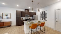 This well-appointed single-floor residence showcases a thoughtfully designed open-concept layout that seamlessly connects the kitchen and living areas. The kitchen is a standout feature, boasting rich dark-wood cabinetry that reaches from floor to ceiling, offering generous storage. A central island with striking granite countertops serves as both a prep area and a casual dining spot, accented by two warm amber leather barstools and illuminated by a pair of brushed nickel industrial-style pendant lights. Stainless steel appliances, including a refrigerator, range, and microwave, complete this functional culinary space. Light wood-tone flooring flows throughout, tying the spaces together with a clean, contemporary aesthetic. The open living area is bright and airy, with recessed lighting and crisp white walls creating a welcoming atmosphere. The bedroom offers a serene retreat, featuring dual windows dressed with sheer linen curtains that invite soft natural light and views of surrounding greenery. A walk-in closet provides convenient storage. The bathroom mirrors the home's refined style with a granite vanity countertop, dark wood cabinetry, and a large mirror with bright overhead lighting, making for a polished and practical space.