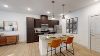 This well-appointed single-floor residence showcases a thoughtfully designed open-concept layout that seamlessly connects the kitchen and living areas. The kitchen is a standout feature, boasting rich dark-wood cabinetry that reaches from floor to ceiling, offering generous storage. A central island with striking granite countertops serves as both a prep area and a casual dining spot, accented by two warm amber leather barstools and illuminated by a pair of brushed nickel industrial-style pendant lights. Stainless steel appliances, including a refrigerator, range, and microwave, complete this functional culinary space. Light wood-tone flooring flows throughout, tying the spaces together with a clean, contemporary aesthetic. The open living area is bright and airy, with recessed lighting and crisp white walls creating a welcoming atmosphere. The bedroom offers a serene retreat, featuring dual windows dressed with sheer linen curtains that invite soft natural light and views of surrounding greenery. A walk-in closet provides convenient storage. The bathroom mirrors the home's refined style with a granite vanity countertop, dark wood cabinetry, and a large mirror with bright overhead lighting, making for a polished and practical space.