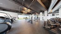 This expansive fitness facility offers a comprehensive workout environment across a single, open floor plan. The space is defined by its industrial-style architecture, featuring exposed ductwork, structural columns, and suspended linear lighting against a dark ceiling — creating a focused, high-energy atmosphere.

The main workout floor is surfaced with durable rubber flooring and is divided into clearly distinct training zones. A prominent artificial turf runway runs along one side of the facility, providing ample space for functional training, agility drills, and dynamic movement exercises. Adjacent to the turf, a Jacob's Ladder climber and stair-stepper machine add to the functional training offerings.

The strength training area is well-appointed with multiple adjustable benches, a full rack of dumbbells, barbells with weight plates, a cable machine system, and a dedicated squat/bench press station — all complemented by floor-to-ceiling mirrors that enhance the sense of depth. Cardio equipment including elliptical machines and a rowing machine is also available throughout the space.

Large windows along one wall allow natural light to fill the space while offering views of the surrounding cityscape. A separate lounge area with light hardwood flooring, seating, and a high-top table provides a comfortable space for rest or social interaction, rounding out this well-rounded fitness facility.