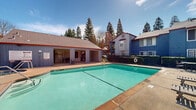 Located at 7849 Sunrise Blvd in Citrus Heights, California, this outdoor communal pool area offers a well-maintained and inviting recreational environment. The centerpiece is a generously sized swimming pool with clear turquoise water, accessible via built-in handrail entries and surrounded by a wide concrete pool deck. Adjacent to the pool is a covered patio structure with an open-air design, featuring exposed beam ceilings and ample space for seating and gathering — ideal for relaxing in the shade during warm California days. Poolside lounge chairs are positioned throughout the deck area, providing comfortable spots to unwind. A security fence encloses the pool area, ensuring a safe and private environment. The surrounding landscape is enriched by mature tall trees, including prominent pines that frame the sky and contribute to a serene, park-like atmosphere. The multi-story residential buildings visible in the background feature a distinctive architectural style with gray and blue vertical board siding, red trim accents, pitched rooflines, and circular window details. The combination of the sparkling pool, shaded lounge areas, and lush natural surroundings makes this outdoor space a functional and appealing community amenity in the heart of Citrus Heights.