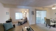 This single-floor living space offers a comfortable and well-lit environment designed for relaxed everyday living. The living area is finished with soft, wall-to-wall carpeting that adds warmth underfoot, while crisp white walls provide a clean, neutral backdrop throughout. A textured ceiling adds subtle visual interest to the room.

Natural light is a defining feature of this space, with a wide horizontal window allowing views of the surrounding greenery outside. A sliding glass door fitted with vertical blinds offers direct access to an outdoor area, further enhancing the connection to the exterior and allowing additional light to filter in. Both window treatments provide practical light control and privacy.

The layout of the living area is open and versatile, providing ample room for seating arrangements. A dedicated lounge corner, visible near the window, creates an inviting nook ideal for reading or relaxing. The overall ambiance is calm and understated, with a neutral color palette that lends itself well to a variety of furnishing styles. The space balances functionality with a light, airy feel, making it an approachable and livable environment.