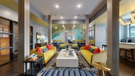 Located at 4206 Simcoe Ln in Louisville, KY, this single-floor community clubhouse is a boldly designed social hub that blends contemporary style with a lively, welcoming atmosphere. The main lounge serves as the centerpiece, featuring striking yellow sofas accented with orange and teal throw pillows, a large abstract mural in muted aquas and greens, and structural columns that frame the open layout. Rich wood-look tile flooring runs throughout, adding warmth and cohesion across the space. A secondary lounge area offers a more relaxed setting, anchored by a deep navy sofa, a glossy dark tile accent wall with a wall-mounted television, and an assortment of seating in complementary colors. Decorative geometric wall art adds a modern touch to this zone. The game room provides a recreational retreat, complete with a billiard table, high-top bistro tables, and curated abstract artwork adorning the walls. A dedicated nook features a teal channel-tufted banquette, a round pedestal table, coral ottomans, and framed artwork, offering an intimate setting for conversation. Large windows and glass doors throughout the space invite abundant natural light and provide views of the surrounding outdoor areas. A kitchenette area with dark cabinetry and countertop space rounds out the amenities, while access doors lead directly to exterior courtyards, seamlessly connecting indoor and outdoor living.