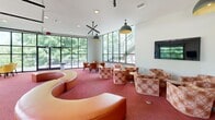 Located at 2201 Jacobs Rd in Seneca, South Carolina, this single-floor clubhouse amenity space at Epoch Student Living offers a generously sized, thoughtfully designed environment built for socializing and community engagement. The entrance features floor-to-ceiling glass panels that flood the foyer with natural light, complemented by wood-look flooring, a branded welcome mat, and a tan leather bench for a polished first impression.

The main lounge area is anchored by a striking curved leather sectional sofa and an assortment of upholstered armchairs arranged beneath bold pendant light fixtures. Large windows and glass doors open to an outdoor terrace, creating a seamless indoor-outdoor connection surrounded by lush tree views. The red-toned carpet adds warmth and vibrancy throughout the gathering zones.

A standout feature is the dual glass-panel garage doors, which open fully to extend the interior into an al fresco seating area furnished with yellow accent chairs and round tables — perfect for casual meetings or relaxed lounging. Wall-mounted televisions are positioned throughout for entertainment.

A bar-style kitchen counter with a warm wood butcher-block top, metal bar stools, and pendant lighting serves as a central hub, equipped with small appliances for residents' convenience. The overall design blends contemporary architecture with inviting, colorful furnishings to create a dynamic and functional communal space.