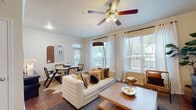This single-floor residence is thoughtfully designed with an open-concept layout that seamlessly connects the main living areas. The living room serves as the central gathering space, featuring rich hardwood-style flooring, crown molding, a ceiling fan with integrated lighting, and large windows dressed with sheer white curtains and blinds that invite generous natural light and views of lush outdoor greenery. The space accommodates both a lounge area and a dedicated dining nook, making it highly functional.

The kitchen is well-appointed with dark espresso cabinetry, granite countertops, stainless steel appliances including a refrigerator, microwave, and range, and a peninsula-style breakfast bar that opens to the main living area. Track lighting above the kitchen adds focused illumination to the workspace.

The bedroom continues the warm aesthetic with plush carpeting, a ceiling fan, and large windows providing ample natural light. The room offers comfortable proportions suitable for a full furniture arrangement.

The bathroom features wood-style flooring, a full tub-and-shower combination with classic white tile surround, a granite-topped vanity with dark cabinetry, and clean, neutral finishes throughout, completing this well-balanced and cohesive residence.