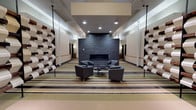 This thoughtfully designed single-floor space offers a series of interconnected communal areas that blend contemporary aesthetics with functional comfort. The layout is anchored by a striking central corridor featuring a floor-to-ceiling dark slate fireplace wall, flanked by clusters of charcoal upholstered armchairs arranged around a circular coffee table — ideal for relaxed conversation or quiet reflection.

Throughout the space, bold horizontal striped flooring in tones of sage green, tan, charcoal, and cream creates a strong visual rhythm that ties the areas together. A defining architectural feature is the recurring sculptural wall installation — a grid of interlocking, wave-shaped panels crafted from light-painted wood and rich dark reclaimed timber — adding texture, depth, and artistic character to multiple zones.

One area is dedicated to casual dining, outfitted with sleek black and lime-green chairs surrounding dark tabletops, complemented by a bar-height counter with black stools. Pendant lighting in warm tones enhances the inviting atmosphere. A separate lounge area features a full sofa arrangement, additional armchairs, a wall-mounted television, and an entertainment unit.

A historical mural depicting a mill scene adds a meaningful nod to local heritage, while recessed and pendant lighting throughout ensures each zone is well-illuminated. The overall ambiance is polished, welcoming, and purposefully designed for community gathering.