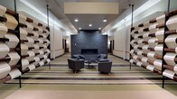 This thoughtfully designed single-floor space offers a series of interconnected communal areas that blend contemporary aesthetics with functional comfort. The layout is anchored by a striking central corridor featuring a floor-to-ceiling dark slate fireplace wall, flanked by clusters of charcoal upholstered armchairs arranged around a circular coffee table — ideal for relaxed conversation or quiet reflection.

Throughout the space, bold horizontal striped flooring in tones of sage green, tan, charcoal, and cream creates a strong visual rhythm that ties the areas together. A defining architectural feature is the recurring sculptural wall installation — a grid of interlocking, wave-shaped panels crafted from light-painted wood and rich dark reclaimed timber — adding texture, depth, and artistic character to multiple zones.

One area is dedicated to casual dining, outfitted with sleek black and lime-green chairs surrounding dark tabletops, complemented by a bar-height counter with black stools. Pendant lighting in warm tones enhances the inviting atmosphere. A separate lounge area features a full sofa arrangement, additional armchairs, a wall-mounted television, and an entertainment unit.

A historical mural depicting a mill scene adds a meaningful nod to local heritage, while recessed and pendant lighting throughout ensures each zone is well-illuminated. The overall ambiance is polished, welcoming, and purposefully designed for community gathering.