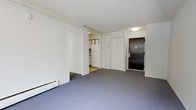 This single-floor unit at Stoneycrest Tower offers a straightforward, functional layout designed to maximize the use of available space. The main living area is finished with low-pile blue-grey carpeting that extends throughout, providing a neutral foundation. White walls and a flat white ceiling keep the space feeling bright and clean. A baseboard heater runs along the lower wall, serving as the primary heat source for the room. A set of white double closet doors provides convenient built-in storage, while a dark-stained entry door with a standard hardware set marks the main entrance. A flush-mount ceiling light fixture near the entry ensures adequate illumination in that zone. An open doorway leads directly into the kitchen, which features white cabinetry, a white refrigerator, and light-toned flooring, distinguishing it from the carpeted living area. The kitchen appears compact but functional, with cabinetry visible along the walls. Overall, the unit presents a clean, no-frills interior with practical features suited for everyday living.