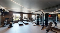 Located at 1700-1706 N Howard St in Philadelphia's 19122 zip code, this contemporary fitness center is thoughtfully designed to support a comprehensive workout experience across a single floor. The space is defined by a striking dark color palette, with charcoal walls and ceilings that create a focused, high-energy atmosphere. A standout architectural feature is the linear LED lighting system, with geometric strips of light running diagonally across the ceiling, providing both functional illumination and a modern aesthetic appeal.

The cardio zone is well-equipped with multiple treadmills, a rowing machine, an elliptical trainer, and a stationary bike, all positioned along windows that bring in natural light and views of the surrounding neighborhood. A large floor-to-ceiling mirror wall visually expands the space while allowing users to monitor their form during exercise.

The strength and functional training area includes a multi-station cable machine, an adjustable bench, a full dumbbell rack, kettlebells, medicine balls, TRX suspension trainers, and a heavy punching bag — offering a diverse range of training options. A dedicated wall-mounted storage rack organizes accessories neatly. The facility also features a water fountain and rubber-composite flooring throughout, ensuring both safety and durability for all workout types.