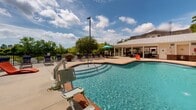 Located at 505 Vorils Ln in Wilmington, NC, this outdoor pool facility offers a well-appointed recreational space designed for both relaxation and leisure. The centerpiece is a generously sized freeform swimming pool with clear blue water, framed by wood-accented coping and surrounded by a broad concrete deck. The pool is equipped with an accessibility lift, ensuring inclusive use for all guests.

The expansive sundeck is furnished with an abundance of lounge chairs and chaise lounges in a variety of styles, providing ample space for sunbathing and socializing. Colorful potted plants and decorative touches add vibrancy to the setting. A pergola structure anchors one corner of the deck, offering a shaded gathering spot, while umbrella-shaded seating clusters provide additional relief from the sun.

A single-story amenity building with a metal roof sits adjacent to the pool and features restroom and facility access. The building's exterior incorporates stone veneer accents, complementing the overall aesthetic of the space.

The entire pool area is enclosed by an iron perimeter fence, with mature trees lining the backdrop, creating a natural, private atmosphere. Lighting poles ensure the space remains functional into the evening hours. The combination of practical amenities and thoughtful design makes this a comfortable and inviting outdoor retreat.