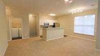 Located at 1000 Henrico Ln in Morrisville, NC, this single-floor residence offers a well-designed open-concept layout that seamlessly connects the kitchen and living areas. The kitchen is a functional centerpiece, featuring white cabinetry, a granite-topped peninsula island, stainless steel appliances including a refrigerator and microwave, and tile backsplash accents that add a clean, polished look. The open flow into the living area creates a generous, airy feel, enhanced by neutral beige carpet that extends throughout the main space. A ceiling fan with a light fixture overhead provides both comfort and illumination, complementing the natural light that filters in through a window along the exterior wall. Track lighting adds additional brightness to the kitchen and dining area. The warm, neutral color palette of soft beige tones on the walls and floors creates a welcoming and versatile ambiance throughout. A hallway visible from the main living area suggests additional private rooms beyond, maintaining a clear distinction between living and personal spaces. Overall, the layout is practical and inviting, making it well-suited for everyday living.