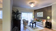 This single-story space showcases a thoughtfully designed living area that balances comfort and style. The room features crown molding along the ceiling perimeter, adding an elegant architectural detail that frames the space with a polished finish. A decorative pendant light fixture serves as a focal point overhead, providing warm ambient lighting that complements the room's inviting atmosphere.

One wall is painted in a rich, deep brown tone, creating a bold contrast against the lighter surrounding walls and serving as a striking backdrop for a framed botanical art piece. Large sliding glass doors fitted with vertical blinds allow abundant natural light to flow into the space while offering access to an outdoor area, enhancing the sense of openness.

The living area is furnished with a neutral-toned sofa accented with colorful throw pillows in green, orange, and warm earth tones, alongside a wicker accent chair that adds texture and casual elegance. A dark wood coffee table anchors a plush patterned area rug over hardwood-style flooring. A tall indoor plant in the corner introduces a fresh, natural element, contributing to the warm and welcoming character of the space.