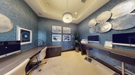 This well-appointed business center room offers a thoughtfully designed workspace with a distinctive aesthetic. The walls are fully clad in rich, textured blue wallcovering that lends the space a bold and professional character. A coffered tray ceiling with crisp crown molding adds architectural depth, while a central drum pendant light fixture provides warm, focused illumination throughout the room. Two dark-toned built-in workstations line the walls, each offering generous surface area suitable for computing and administrative tasks, with an iMac computer and printer readily accessible. Ergonomic rolling office chairs accompany the desks, ensuring comfort during extended use. A low dark-wood storage cabinet anchors one corner, providing additional organizational capacity. Decorative elements elevate the room's ambiance, including a curated set of four framed art prints arranged in a grid, a sculptural metallic wall installation of overlapping disc forms, and a tall potted bamboo plant that introduces a touch of greenery. Light-toned carpet flooring completes the refined look. A glass-paneled door with a flowing gray curtain connects the room to an adjacent lounge or common area, allowing for both privacy and easy access.
