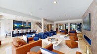 Located at 141 Danbury, this thoughtfully designed communal amenity space offers a generous, open-plan layout across a single floor. The interior is defined by clean white ceilings with recessed lighting, light hardwood and herringbone-patterned tile flooring, and an airy, welcoming atmosphere throughout.

The lounge area is anchored by a striking built-in electric fireplace set against a textured geometric tile feature wall, flanked by warm wood shelving units displaying curated décor and greenery. A large wall-mounted display screen adds a media-friendly focal point. Seating is arranged in distinct conversation zones, mixing bold cobalt-blue tufted sofas with cognac leather armchairs for a vibrant yet cohesive color palette.

A sleek staircase with open metal railings provides visual interest and connects to upper levels. The kitchen and dining area features a large white quartz island with built-in cooktop and bar seating for multiple guests, accented by brass pendant lighting and a dramatic navy hexagonal tile backsplash with dual mounted screens.

Floor-to-ceiling sliding glass doors line one side of the space, flooding the interior with natural light and offering direct access to an outdoor terrace. Lush potted trees placed throughout soften the contemporary design and reinforce an inviting, resort-like ambiance.