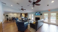 This single-floor community space is thoughtfully designed for both comfort and functionality, offering a seamless flow between its key areas. The expansive main living room is anchored by a classic fireplace and a wall-mounted television, with vaulted ceilings, dual ceiling fans, and recessed lighting creating an airy, inviting atmosphere. Rich hardwood flooring runs throughout, complementing the light blue and neutral gray wall tones. Generous seating, including leather sofas and tufted accent chairs, makes the space ideal for gathering. French doors open directly to an outdoor pool area, flooding the interior with natural light and providing effortless indoor-outdoor access. Adjacent to the living room, the open-concept kitchen features dark espresso cabinetry, granite countertops, black appliances, and a peninsula bar, offering both ample prep space and a casual serving area. A separate dining area accommodates a large stone-top table with upholstered seating, decorated with elegant wall accents and crown molding. The overall ambiance is modern yet comfortable, blending practical amenities with stylish finishes suited for community events, social gatherings, or everyday use.