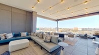 This contemporary elevated lounge is a thoughtfully designed indoor-outdoor amenity space that blends comfort with striking panoramic views of the surrounding cityscape. The space is anchored by large-format porcelain tile flooring that extends seamlessly from the interior lounge to the open terrace, reinforcing the fluid connection between the two zones.

The interior lounge area features a curated mix of seating, including a light gray sectional sofa and a teal accent sofa accented with geometric throw pillows, arranged to encourage relaxed social interaction. A wall-mounted television and a large potted ornamental grass provide decorative focal points against the clean, panel-clad walls finished in muted gray tones.

Overhead, a track lighting system with gold brass fixtures runs across the white ceiling, delivering warm ambient illumination that complements the natural daylight flooding in through the expansive glass wall. This floor-to-ceiling glass panel system opens fully to the outdoor terrace, creating an unobstructed transition to the open-air seating area beyond.

The terrace extends the lounge outward with additional modular seating, round bistro tables, and side tables, all positioned to take full advantage of the elevated views. A glass perimeter railing preserves sightlines to the horizon, while structured planters add a natural design element. The overall ambiance is refined, airy, and modern.