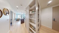 This thoughtfully designed single-floor residence offers a seamless blend of modern aesthetics and functional living. Light-toned hardwood flooring flows throughout the space, unifying each area with a warm, cohesive feel. A built-in shelving unit anchors the entryway, providing both storage and a stylish display area before opening into the heart of the home.

The kitchen is a standout, featuring flat-front cabinetry in a soft white and natural wood-grain finish, under-cabinet lighting, a textured white tile backsplash, and stainless steel appliances including a full-size range and refrigerator. A central island adds prep space and visual continuity.

The living area is bathed in natural light through expansive floor-to-ceiling windows with bold dark frames, offering views of the surrounding urban streetscape. The open layout encourages effortless flow between the living and dining zones.

The bedroom is a calm and comfortable retreat, featuring a large window that frames city views while allowing ample daylight. Clean white walls and light flooring contribute to the serene atmosphere.

The bathroom is finished with large-format neutral floor tiles and a floating double vanity in a warm wood tone, topped with a white countertop and paired with dual rounded black-framed mirrors and sleek linear lighting above.