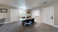 Located at 1175 Lake Blvd in Davis, California, this single-floor office space offers a well-organized and versatile environment designed to support both collaborative meetings and focused individual work. The room is divided into two functional zones: a conference area and a dedicated workstation section.

The conference area is anchored by a large, dark oval table surrounded by ergonomic task chairs, making it well-suited for team meetings or client presentations. A full-sized whiteboard mounted on the wall provides an ideal surface for brainstorming and visual communication. A potted plant adds a touch of greenery, softening the professional atmosphere. Natural light filters in through a large window fitted with vertical blinds, complemented by a ceiling fan with a light fixture and a track lighting system that ensures consistent illumination throughout.

The workstation area features a long, wall-mounted built-in desk that comfortably accommodates multiple users simultaneously. Each station is equipped with monitors and office supplies, supported by adjustable task chairs. Track lighting above the desk provides focused task lighting, while a bulletin board on the adjacent wall keeps reference materials accessible.

The space features neutral-toned walls, textured carpet flooring, and clean white trim throughout, creating a calm, professional ambiance conducive to productivity.