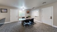 Located at 1175 Lake Blvd in Davis, California, this single-floor office space offers a well-organized and versatile environment designed to support both collaborative meetings and focused individual work. The room is divided into two functional zones: a conference area and a dedicated workstation section.

The conference area is anchored by a large, dark oval table surrounded by ergonomic task chairs, making it well-suited for team meetings or client presentations. A full-sized whiteboard mounted on the wall provides an ideal surface for brainstorming and visual communication. A potted plant adds a touch of greenery, softening the professional atmosphere. Natural light filters in through a large window fitted with vertical blinds, complemented by a ceiling fan with a light fixture and a track lighting system that ensures consistent illumination throughout.

The workstation area features a long, wall-mounted built-in desk that comfortably accommodates multiple users simultaneously. Each station is equipped with monitors and office supplies, supported by adjustable task chairs. Track lighting above the desk provides focused task lighting, while a bulletin board on the adjacent wall keeps reference materials accessible.

The space features neutral-toned walls, textured carpet flooring, and clean white trim throughout, creating a calm, professional ambiance conducive to productivity.