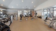 This fitness center is a thoughtfully designed, single-floor facility offering a wide range of workout options across several distinct zones. The entrance features large glass doors and floor-to-ceiling glazing that floods the entryway with natural light, creating a welcoming and open arrival experience.

The cardio area houses a row of modern treadmills positioned in front of windows overlooking the surrounding neighborhood, allowing users to exercise with an outdoor view. Adjacent to this is a dedicated cardio machine zone lined with elliptical trainers, complemented by a dumbbell rack and a wall-mounted flat-screen TV for entertainment during workouts.

The strength training area is expansive, featuring high vaulted ceilings and recessed lighting that enhance the sense of openness. It is outfitted with a variety of cable machines, weight benches, functional trainers, and specialty resistance equipment arranged across durable rubber flooring, with large mirrors along the walls to support proper form.

A separate studio room provides a dedicated space for flexibility, balance, and group-style training. It features hardwood-style flooring, full-length mirrors, a large display screen, and an assortment of stability balls and balance trainers. Windows throughout the studio bring in natural light and maintain an airy atmosphere. The overall facility presents a clean, modern aesthetic with neutral tones and practical, performance-oriented design.