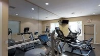 This fitness center is designed to provide a comprehensive cardio-focused workout environment within a single-level layout. The room features floor-to-ceiling mirrors along one wall, which visually expand the space and allow users to monitor their form during exercise. Recessed lighting in the smooth ceiling ensures the room is evenly and brightly illuminated throughout.

The equipment lineup includes treadmills, elliptical cross-trainers, and stationary bikes, offering a range of low-impact and moderate-intensity cardio options. Each machine is positioned to allow comfortable use without overcrowding. A wall-mounted flat-screen television is installed near the window, providing entertainment during workouts.

Natural light filters into the room through a window overlooking an outdoor area, contributing to an open and welcoming atmosphere. The neutral beige walls and light-toned ceiling complement the clean, no-frills aesthetic of the space. Ventilation is provided through ceiling vents, supporting air circulation during active use. The overall design is practical and user-friendly, prioritizing functionality and ease of movement throughout the fitness area.