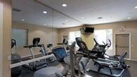This fitness center is designed to provide a comprehensive cardio-focused workout environment within a single-level layout. The room features floor-to-ceiling mirrors along one wall, which visually expand the space and allow users to monitor their form during exercise. Recessed lighting in the smooth ceiling ensures the room is evenly and brightly illuminated throughout.

The equipment lineup includes treadmills, elliptical cross-trainers, and stationary bikes, offering a range of low-impact and moderate-intensity cardio options. Each machine is positioned to allow comfortable use without overcrowding. A wall-mounted flat-screen television is installed near the window, providing entertainment during workouts.

Natural light filters into the room through a window overlooking an outdoor area, contributing to an open and welcoming atmosphere. The neutral beige walls and light-toned ceiling complement the clean, no-frills aesthetic of the space. Ventilation is provided through ceiling vents, supporting air circulation during active use. The overall design is practical and user-friendly, prioritizing functionality and ease of movement throughout the fitness area.