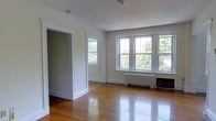 This single-floor space showcases a well-maintained interior defined by warm hardwood floors that run throughout the main living area, their polished surface reflecting ample natural light. The room is anchored by a bank of multi-pane double-hung windows along one wall, offering expansive views of surrounding greenery and flooding the interior with bright, diffused daylight. An additional single window on an adjacent wall further enhances the sense of openness and cross-lighting within the space. The walls are finished in a soft, neutral tone that complements the warm wood flooring and contributes to a calm, understated ambiance. Classic white trim and baseboards frame the doorways and walls, adding a clean architectural detail consistent with traditional construction. A radiator beneath the main window grouping indicates a hot-water heating system, a hallmark of older, well-built residential construction. The open doorway visible in the space leads to an adjoining room, suggesting a functional, connected layout. Overall, the space presents a bright, classic character with quality original finishes and generous natural light.