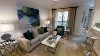This single-floor space showcases a thoughtfully designed living area that balances elegance with comfort. The room is anchored by a neutral-toned sofa dressed with teal and olive accent pillows, complemented by a deep green accent chair that adds a rich contrast to the light, warm color palette throughout. Crown molding lines the ceiling, lending the space a refined architectural detail that elevates the overall aesthetic.

A large, bold abstract painting in shades of teal, navy, and green serves as a dramatic focal point on one wall, tying the color scheme together cohesively. A glass-topped coffee table with a metallic frame sits at the center of the seating arrangement, adding a contemporary touch while maintaining an open, airy feel.

Natural light filters into the room through a generously sized window dressed with layered blinds and floor-length draping curtains in a soft neutral tone, offering both style and light control. The carpeted flooring in a warm beige tone contributes to the cozy ambiance of the space. Side tables with slim metallic legs complete the arrangement, providing functional surfaces without overwhelming the room's balanced, well-curated design.