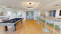 Located at 116 NE Third St in Pompano Beach, Florida, this single-floor amenity lounge is a generously sized, thoughtfully designed communal space that balances recreation and relaxation. The interior features light hardwood flooring throughout, crisp white walls, and a clean modern aesthetic accented by colorful artwork.

The game zone is a focal point of the space, housing a full-size billiards table beneath a striking circular pendant light with a coffered ceiling detail, complemented by a foosball table nearby. High-top bar tables with green-cushioned stools provide casual seating in this area, ideal for social gatherings.

The lounge area transitions seamlessly into a comfortable living space furnished with a large sectional sofa, accent chairs in soft blue and grey tones, and a patterned area rug. A wall-mounted large-screen television anchors one end of the room, surrounded by multiple oversized windows that flood the space with natural light and frame panoramic views of the surrounding Pompano Beach skyline and lush greenery.

Floor-to-ceiling windows along one wall overlook an outdoor pool area with vibrant plantings, enhancing the bright, airy atmosphere. Recessed lighting and flush drum pendant fixtures provide ample illumination throughout. The overall ambiance is contemporary, inviting, and resort-inspired, making it an ideal multipurpose amenity space.