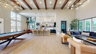 Located at 3755 E McKinney St in Denton, Texas, this single-floor community clubhouse offers a generously sized, open-concept layout designed for both socializing and relaxation. The space is defined by its dramatic soaring ceilings adorned with rich, dark-stained exposed wood beams, creating a warm yet contemporary architectural statement throughout.

The welcoming reception area features shiplap wall paneling, a warm wood desk, and a grand circular chandelier that sets an inviting tone from entry. Large multi-pane windows and glass doors flood the interior with abundant natural light, offering views of the outdoor pool and surrounding landscaping.

The expansive main living area is anchored by a stylish billiards table and a sculptural geometric chandelier, with multiple distinct seating zones furnished with plush sofas, leather accent chairs, and wood coffee tables. Light-toned hardwood-style flooring runs seamlessly throughout, enhancing the sense of openness.

A vibrant kitchen and dining nook adds functionality and artistic flair, featuring teal cabinetry, a white quartz island, pendant lighting, and a bold geometric mural that energizes the space. An intimate lounge area with a wall-mounted TV and pool-facing floor-to-ceiling windows rounds out this well-appointed clubhouse, making it a dynamic hub for community life.
