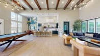 Located at 3755 E McKinney St in Denton, Texas, this single-floor community clubhouse offers a generously sized, open-concept layout designed for both socializing and relaxation. The space is defined by its dramatic soaring ceilings adorned with rich, dark-stained exposed wood beams, creating a warm yet contemporary architectural statement throughout.

The welcoming reception area features shiplap wall paneling, a warm wood desk, and a grand circular chandelier that sets an inviting tone from entry. Large multi-pane windows and glass doors flood the interior with abundant natural light, offering views of the outdoor pool and surrounding landscaping.

The expansive main living area is anchored by a stylish billiards table and a sculptural geometric chandelier, with multiple distinct seating zones furnished with plush sofas, leather accent chairs, and wood coffee tables. Light-toned hardwood-style flooring runs seamlessly throughout, enhancing the sense of openness.

A vibrant kitchen and dining nook adds functionality and artistic flair, featuring teal cabinetry, a white quartz island, pendant lighting, and a bold geometric mural that energizes the space. An intimate lounge area with a wall-mounted TV and pool-facing floor-to-ceiling windows rounds out this well-appointed clubhouse, making it a dynamic hub for community life.