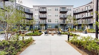 Located at 4455 Condor Dr in Hilliard, Ohio, this outdoor courtyard is a thoughtfully designed communal space nestled within a modern multi-story residential building. The courtyard is enclosed on multiple sides by the building's contemporary façade, featuring grey paneling, large windows, and private balconies with black metal railings and warm wood-accented overhangs that lend an inviting architectural character.

At ground level, the space is defined by a network of concrete pathways weaving through richly planted garden beds filled with seasonal shrubs, ornamental grasses, and young trees. String lights suspended overhead add a warm ambiance, particularly in the evening. Seating is distributed throughout in varied forms — woven egg chairs, cushioned lounge chairs, and grouped conversation sets — offering residents multiple areas to relax or gather.

Recreational amenities include a hammock installation and a cornhole board, contributing to the courtyard's social atmosphere. A stone-accented fire feature anchors one seating zone, while a covered passageway adorned with a bold, large-scale artistic mural provides a striking visual element and connects the courtyard to the surrounding area. Landscape lighting and architectural bollards further enhance the space after dark. The overall design balances greenery, art, and functional outdoor living in a cohesive and welcoming environment.