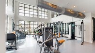 Located at 445 51st St in Wilmington, NC, this contemporary two-floor fitness center offers a well-appointed and thoughtfully designed workout facility spanning two generously sized levels. The ground floor serves as the primary strength training area, distinguished by soaring double-height ceilings and expansive floor-to-ceiling windows framed in sleek black steel. These large-pane windows flood the space with abundant natural light while providing open views of the surrounding landscape. The strength floor is equipped with a comprehensive selection of resistance machines, free weights, benches, cable systems, and functional training rigs, all set on durable, easy-to-clean flooring. An artistic mural accent wall adds visual interest near the entry area, which features a clean, open layout with a central staircase outfitted with industrial-style mesh railings leading to the upper level. The second floor houses a dedicated cardio zone featuring a row of treadmills and elliptical machines positioned alongside windows that overlook the exterior grounds. Exposed structural beams and recessed lighting lend this level an industrial-modern character. Separated from the main floor, a private cycling studio features smooth hardwood flooring, wall-to-wall mirrors, mounted displays, and Peloton-style stationary bikes, creating an immersive, studio-quality spin experience. Together, the two floors deliver a comprehensive, premium fitness environment.