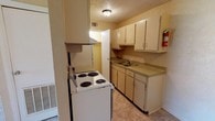 This single-floor unit at 3630 Rainey Rd in Jackson, Mississippi offers a straightforward and functional layout designed for practical everyday living. The galley-style kitchen is efficiently arranged with cream-colored cabinetry lining both walls, providing ample storage space. It is equipped with an electric coil range with overhead ventilation, a refrigerator, a double-basin sink, and continuous countertop workspace. Tile flooring runs throughout the kitchen, which also connects to a closet or utility area through a doorway at the rear. The living area features a combination of tile and carpet flooring, with textured walls painted in a warm neutral tone and a textured ceiling throughout the unit. Natural light enters through windows with horizontal blinds. The bedroom is carpeted and offers a quiet, open space with a window bringing in additional natural light. The full bathroom includes a pedestal-style vanity with a mirror above, a toilet, and a full-size bathtub with tiled surround and a tile accent bar. Overhead lighting and a textured ceiling are consistent throughout. The unit's neutral color palette and practical design make it a functional space well-suited for comfortable single-floor living.