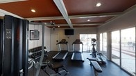 This fitness center offers a comprehensive workout environment designed to support a range of exercise routines. The space is anchored by a striking coffered ceiling finished in a rich, deep red tone, accented by recessed lighting that provides even illumination throughout the room. A wall-mounted flat-screen TV keeps members entertained during cardio sessions.

The cardio area features multiple treadmills and an elliptical trainer, all positioned to take advantage of large floor-to-ceiling windows along one wall. These windows flood the space with natural light and offer views of the outdoor surroundings, creating an open, energizing atmosphere despite the interior setting.

Along the opposite wall, a well-stocked dumbbell rack holds an extensive range of free weights, accompanied by a weight bench, making the space suitable for both beginners and experienced fitness enthusiasts. A cable machine further expands strength training options. The dark rubber flooring throughout provides durability, comfort underfoot, and safety during workouts. Decorative framed artwork adorns the walls, adding a polished, welcoming touch to the functional environment. Overall, this fitness center balances practical amenities with thoughtful design elements to create an inviting and motivating workout space.