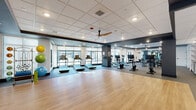 Located at 5801 Sanderson St NW in Huntsville, Alabama, this fitness center offers a generously sized, single-floor layout designed to accommodate a full range of exercise activities. The space is thoughtfully divided into distinct functional zones, each finished with purpose-appropriate flooring — resilient rubber flooring in the equipment areas and warm hardwood-style flooring in the open studio zone.

The cardio section features a row of treadmills, elliptical machines, and stationary bikes positioned along large floor-to-ceiling windows that invite abundant natural light and provide views of the surrounding outdoor environment. The strength training area is equipped with a comprehensive dumbbell rack, weight benches, and a cable machine, complemented by full-length mirrors that enhance the sense of space. Ceiling fans and recessed lighting ensure a comfortable, well-lit environment throughout.

The open studio area provides an expansive, unobstructed floor ideal for group fitness classes, stretching, or functional training, with stability balls and accessories readily available. A welcoming entry area features a built-in storage cubby wall, a check-in counter with cabinetry, and glass-paneled doors framed by a stone accent column. The design palette of dark trim, white walls, and gray tones gives the entire fitness center a clean, contemporary aesthetic.