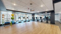 Located at 5801 Sanderson St NW in Huntsville, Alabama, this fitness center offers a generously sized, single-floor layout designed to accommodate a full range of exercise activities. The space is thoughtfully divided into distinct functional zones, each finished with purpose-appropriate flooring — resilient rubber flooring in the equipment areas and warm hardwood-style flooring in the open studio zone.

The cardio section features a row of treadmills, elliptical machines, and stationary bikes positioned along large floor-to-ceiling windows that invite abundant natural light and provide views of the surrounding outdoor environment. The strength training area is equipped with a comprehensive dumbbell rack, weight benches, and a cable machine, complemented by full-length mirrors that enhance the sense of space. Ceiling fans and recessed lighting ensure a comfortable, well-lit environment throughout.

The open studio area provides an expansive, unobstructed floor ideal for group fitness classes, stretching, or functional training, with stability balls and accessories readily available. A welcoming entry area features a built-in storage cubby wall, a check-in counter with cabinetry, and glass-paneled doors framed by a stone accent column. The design palette of dark trim, white walls, and gray tones gives the entire fitness center a clean, contemporary aesthetic.