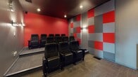 This private screening room is purpose-built to deliver a true cinematic experience in an intimate setting. The room is defined by its bold design choices, featuring striking red accent walls that contrast sharply with a dark, matte ceiling and neutral grey tones throughout. Multiple rows of black leather theater-style seats are arranged in a tiered, stadium configuration, ensuring clear sightlines from every position. Each seat is equipped with built-in armrests and cup holders, emphasizing comfort during extended viewing sessions. The side and rear walls are adorned with a checkerboard pattern of red and grey acoustic panels, which serve both a decorative and functional purpose by managing sound quality within the room. Recessed ceiling lighting and wall-mounted sconces provide controlled, dimmable illumination suited to a screening environment. A dedicated control area near the entrance features a wall-mounted display screen, an AV equipment console, and movie posters that reinforce the cinematic theme. The carpeted flooring throughout adds to the acoustic dampening of the space. Access is provided through a glass-paneled door, and a wall clock is mounted near the entry for practical use. Overall, this screening room offers a well-appointed, professional-grade environment for private film viewing or presentations.