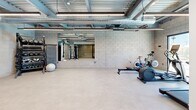 Located in Phoenix, AZ, this fitness center offers a single-floor exercise space designed with an industrial-modern aesthetic. The room features exposed ceilings with visible ductwork, metal decking, and linear LED lighting that creates a bright, energetic atmosphere well-suited for physical activity.

The exercise room is comprehensively equipped for a full-body workout. Cardio equipment includes treadmills, a rowing machine, a spin bike, and an elliptical trainer, while the strength training area offers a cable machine with an adjustable bench, a rack of dumbbells and kettlebells in varying weights, and a stability ball. A large wall-mounted mirror enhances the sense of space and supports proper form during exercise, and a flat-screen TV is mounted on the concrete block wall for entertainment.

A standout feature is the large sliding glass window that frames a direct view of an outdoor swimming pool, filling the space with natural light and providing a refreshing visual connection to the exterior. The durable, light-toned flooring is practical and easy to maintain. Entry to the facility is through glass-paneled doors, adjacent to a vending machine area offering convenient refreshments. The concrete block walls and open ceiling lend the space an urban, functional character ideal for focused training.