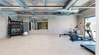 Located in Phoenix, AZ, this fitness center offers a single-floor exercise space designed with an industrial-modern aesthetic. The room features exposed ceilings with visible ductwork, metal decking, and linear LED lighting that creates a bright, energetic atmosphere well-suited for physical activity.

The exercise room is comprehensively equipped for a full-body workout. Cardio equipment includes treadmills, a rowing machine, a spin bike, and an elliptical trainer, while the strength training area offers a cable machine with an adjustable bench, a rack of dumbbells and kettlebells in varying weights, and a stability ball. A large wall-mounted mirror enhances the sense of space and supports proper form during exercise, and a flat-screen TV is mounted on the concrete block wall for entertainment.

A standout feature is the large sliding glass window that frames a direct view of an outdoor swimming pool, filling the space with natural light and providing a refreshing visual connection to the exterior. The durable, light-toned flooring is practical and easy to maintain. Entry to the facility is through glass-paneled doors, adjacent to a vending machine area offering convenient refreshments. The concrete block walls and open ceiling lend the space an urban, functional character ideal for focused training.