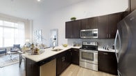 Located at Woodmont Metro at Metuchen Station, this single-floor residence showcases a thoughtfully designed interior with modern finishes throughout. The kitchen serves as a central highlight, featuring rich, dark espresso-toned cabinetry that extends to the upper walls, providing generous storage space. Complementing the cabinetry are crisp white quartz countertops that offer ample prep space, creating a striking contrast against the darker tones. A full suite of stainless steel appliances, including a range with oven, built-in microwave, and dishwasher, adds a polished, contemporary feel. The tile backsplash in a subtle gray stone pattern adds texture and visual interest to the space. Light wood-toned flooring runs throughout, contributing warmth and continuity to the overall design. Bright white walls and recessed lighting enhance the sense of openness, while the layout of the kitchen allows for efficient workflow and functionality. The space reflects a clean, modern aesthetic that balances style with practicality, making it well-suited for everyday living.