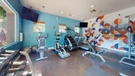 Located at 6233 Dougherty Rd in Dublin, California, this single-floor fitness center is designed to provide a comprehensive and motivating workout environment. The space is defined by bold teal accent walls and a striking, large-scale geometric mural depicting an athletic figure in motion — rendered in a vivid palette of orange, blue, and cream — that serves as an energizing focal point for the room.

The facility is well-equipped with a diverse selection of cardio machines, including multiple treadmills, a StairMaster step climber, an elliptical, and a stationary bike. For strength training, the space features a cable machine, a full rack of dumbbells, and a weight bench, offering a well-rounded workout experience. Multiple wall-mounted flat-screen TVs are positioned throughout the room for entertainment during workouts.

Natural light flows in through several windows, offering views of the surrounding outdoor greenery and streetscape, creating an open and inviting atmosphere. Recessed lighting supplements the natural light, ensuring the space remains bright at all hours. The flooring consists of durable, dark rubberized material suited for high-traffic fitness use. The overall design is clean, modern, and purposefully laid out to maximize functionality while maintaining a visually dynamic and energizing aesthetic.