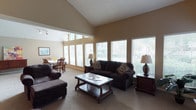 This single-story space showcases an open and airy living area anchored by soaring vaulted ceilings that lend a generous sense of height and volume. The living room is furnished with dark brown upholstered sofas and a matching oversized chair with ottoman, arranged around a classic wooden coffee table with a lower display shelf. Neutral-toned carpet throughout the main area complements the warm beige walls and textured ceiling.

A standout feature of this space is the abundance of natural light, made possible by a generous arrangement of large windows fitted with white horizontal blinds. These windows frame views of lush outdoor greenery, bringing a connection to nature into the interior. Floor lamps with warm-toned shades on either side of the seating area provide additional ambient lighting.

Adjacent to the living room, a dining area is visible, featuring a round table with wooden chairs and additional windows that further enhance the bright, welcoming atmosphere. The overall ambiance is comfortable and relaxed, with a traditional aesthetic achieved through the combination of rich, dark wood furnishings and soft, neutral finishes throughout.