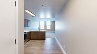 This single-floor unit offers a thoughtfully designed layout that balances functionality and comfort. The kitchen serves as a central hub of the space, featuring rich dark wood cabinetry, a peninsula with a built-in sink, and a full suite of stainless steel appliances including a refrigerator, electric range, over-the-range microwave, and dishwasher. Light wood-look flooring runs throughout the kitchen and entry areas, lending warmth and easy maintenance to the high-traffic zones. The kitchen opens seamlessly toward the living area, creating an inviting, open-concept flow. The bedrooms are carpeted and bathed in natural light through multiple windows fitted with roller shades. One of the larger bedrooms features a wraparound arrangement of windows offering expansive city views, making it a standout feature of the unit. Additional bedrooms are well-proportioned with individual windows providing further urban vistas. The bathroom is clean and functional, equipped with a white vanity with ample cabinet storage, a medicine cabinet mirror, and a full tub-shower combination. Neutral, light-toned walls throughout the unit create a bright and airy ambiance, complementing the modern finishes and making the space feel open and move-in ready.