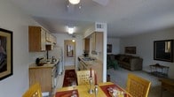 This single-story residence presents an open-concept floor plan that seamlessly integrates the kitchen, dining area, and living room into one cohesive living space. The kitchen is well-appointed with light wood cabinetry extending to the upper walls, offering generous storage. It features a electric range, a refrigerator, and a double sink set into a continuous countertop. A structural column and half-wall subtly define the kitchen from adjacent areas without sacrificing the open feel. The dining area accommodates a sizable table with lattice-back chairs, set with candles and place settings, creating an inviting atmosphere for meals. A decorative area rug anchors the space beneath. The living room, visible beyond the dining area, features neutral carpet and is furnished with a large sectional sofa, framed wall art, and a tall potted plant that adds a natural element. Textured ceilings are consistent throughout, and overhead lighting fixtures illuminate each zone. A hallway visible from the kitchen leads to additional areas of the home, suggesting a practical and well-organized layout throughout.