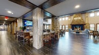 This expansive single-story community space is defined by its rustic lodge-inspired design, blending natural materials with thoughtfully appointed gathering areas. Throughout the space, reclaimed wood-clad columns and dark exposed beam ceilings create a warm, cohesive aesthetic, while dark wood-look tile flooring flows seamlessly from room to room.

The great room serves as a dramatic centerpiece, featuring soaring double-height ceilings, rows of large windows that draw in abundant natural light, and a commanding floor-to-ceiling stone fireplace flanked by seating areas. An elegant chandelier anchors the space above.

Adjacent to the great room, a full kitchen is outfitted with rich wood cabinetry, a stone-topped island with bar seating, stainless steel appliances, a decorative tile backsplash, and pendant lighting — ideal for communal cooking and dining. A dedicated bar area with counter seating and mounted televisions provides an additional social hub.

A separate dining area accommodates multiple table groupings and opens toward a dedicated theater room, complete with tiered seating and a projection screen. Wide corridors lined with reclaimed wood columns and original artwork connect the various areas, maintaining the space's warm, welcoming character throughout.