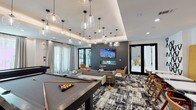 The Social Lounge is a thoughtfully designed, single-floor amenity space that seamlessly blends entertainment, dining, and relaxation into one cohesive environment. The space is divided into several distinct functional zones, each with its own character and purpose.

The game area anchors one side of the lounge, featuring two full-sized billiard tables positioned beneath clusters of modern glass pendant lights that hang from a coffered ceiling. A built-in counter with white bar stools along the perimeter provides additional casual seating. Large windows dressed with sheer drapes allow abundant natural light to fill the space.

Adjacent to the game area, a sophisticated lounge seating zone is defined by a bold, dark accent wall clad in large-format tile, which frames a wall-mounted flat-screen television and a sleek linear fireplace beneath it. Comfortable sofas, armchairs, and a tufted leather ottoman are arranged on a graphic black-and-white area rug, creating an inviting gathering spot.

A dedicated bar and wine storage area features floor-to-ceiling dark cabinetry with wine racks, a mirrored backsplash, under-counter refrigeration, and a statement wood-and-brass high-top table. A separate catering kitchen offers an extended white quartz countertop, stainless steel appliances, a mosaic tile backsplash, and a wall-mounted television, with a small bistro seating nook finished in warm wood paneling nearby.