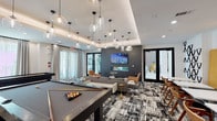 The Social Lounge is a thoughtfully designed, single-floor amenity space that seamlessly blends entertainment, dining, and relaxation into one cohesive environment. The space is divided into several distinct functional zones, each with its own character and purpose.

The game area anchors one side of the lounge, featuring two full-sized billiard tables positioned beneath clusters of modern glass pendant lights that hang from a coffered ceiling. A built-in counter with white bar stools along the perimeter provides additional casual seating. Large windows dressed with sheer drapes allow abundant natural light to fill the space.

Adjacent to the game area, a sophisticated lounge seating zone is defined by a bold, dark accent wall clad in large-format tile, which frames a wall-mounted flat-screen television and a sleek linear fireplace beneath it. Comfortable sofas, armchairs, and a tufted leather ottoman are arranged on a graphic black-and-white area rug, creating an inviting gathering spot.

A dedicated bar and wine storage area features floor-to-ceiling dark cabinetry with wine racks, a mirrored backsplash, under-counter refrigeration, and a statement wood-and-brass high-top table. A separate catering kitchen offers an extended white quartz countertop, stainless steel appliances, a mosaic tile backsplash, and a wall-mounted television, with a small bistro seating nook finished in warm wood paneling nearby.