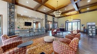 Located at 3918 Atascocita Rd in Humble, TX, this single-floor community lounge and gathering space offers a generously sized, thoughtfully designed interior that blends upscale architectural detail with warm, welcoming ambiance. The centerpiece of the space is its dramatically tall coffered ceiling, framed by bold dark wood beams that add depth and a refined, lodge-inspired character. Elegant tiered chandeliers hang throughout, providing warm ambient lighting that complements the abundant natural light streaming through expansive floor-to-ceiling windows and French doors. Rich hardwood flooring laid in a classic herringbone pattern runs throughout the space, grounding the design with a sense of timeless sophistication. Distinctive latticed decorative columns define separate zones within the open layout, which includes a comfortable living area with plush seating, a bar-height counter area with stools, and casual dining and conversation nooks. Stone tile accent walls and stacked stone details add texture and visual interest. Direct outdoor access through grand arched entry doors connects the interior to the surrounding grounds, offering views of greenery and open skies. The overall atmosphere is inviting and polished, well-suited for socializing and community gatherings.